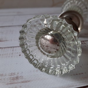 May include: A clear glass door knob featuring a circular, fluted design with a central metallic insert that has a weathered, speckled finish. The knob has a scalloped edge and a reflective, transparent appearance with a visible metal base component.