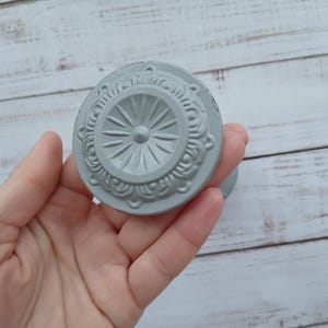 Peut inclure: Un bouton de meuble gris décoratif avec un motif circulaire orné. Le bouton présente un motif central en forme d'éclat d'étoile entouré d'une bordure détaillée et tourbillonnante. Le bouton est tenu sur un fond en bois clair.