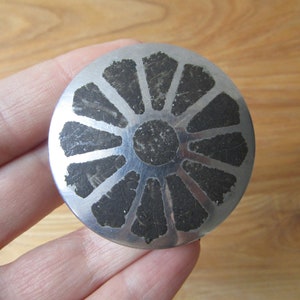 May include: A circular, silver-coloured metal pendant with a radial pattern. The design has dark, textured segments extending from a central circle. The pendant is held in a hand, showing its size and detail.