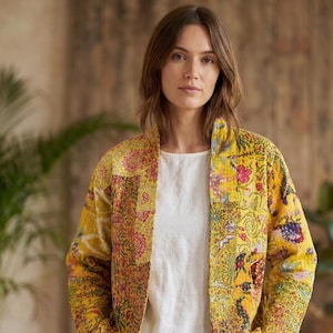 May include: A yellow patchwork jacket with a floral pattern in pink, red, and green. The jacket has a stand-up collar and long sleeves. The person is wearing a white top and light blue jeans. The background is a neutral color.