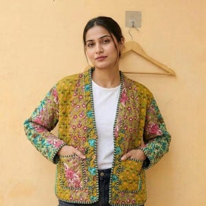 Puede incluir: Chaqueta acolchada con estampado floral en tonos amarillos, rosas y azules. La chaqueta tiene dos bolsillos delanteros y se lleva abierta. Los bordes están ribeteados con una costura verde y blanca contrastante.