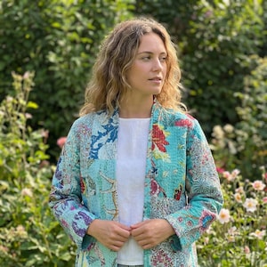 May include: A turquoise patchwork jacket with a floral and bird print. The jacket has a relaxed fit and is worn over a white top and blue jeans. The jacket's design includes various patterns and colors, creating a unique and artistic look.
