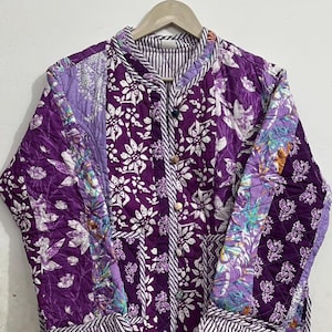 May include: A quilted jacket in shades of purple, featuring floral and paisley patterns. The jacket has a mandarin collar, button closure, and striped cuffs. The jacket is hanging on a wooden hanger.