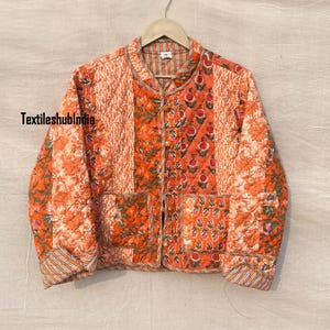 Könnte beinhalten: Eine gesteppte Jacke mit Stehkragen und Knopfleiste. Die Jacke besteht aus einem Patchwork aus verschiedenen Stoffen mit Blumendrucken in Orange- und Weißtönen. Die Jacke hat eine Tasche auf der linken Seite.