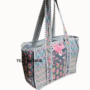 Puede incluir: Un bolso tote acolchado con estampados florales en rosa, gris y turquesa. El bolso tiene asas a rayas y el texto "TEXTILESHUB" en el lateral. El bolso está sostenido por una mano.