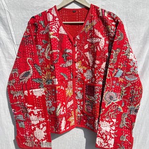 May include: Red quilted jacket with a V-neck and two front pockets. The jacket features a vibrant floral and animal print in white, yellow, and green. The jacket is hanging on a wooden hanger against a white background.