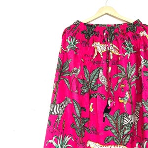 Puede incluir: Falda rosa brillante con estampado de jungla. La tela presenta ilustraciones de cebras, leopardos, loros y follaje verde. La falda está colgada de una percha de madera.