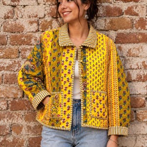 May include: A quilted jacket featuring a vibrant yellow base with multi-coloured floral block print patterns. The garment includes a collared neckline, long sleeves with striped cuffs, two front patch pockets, and a front button closure with matching yellow buttons.