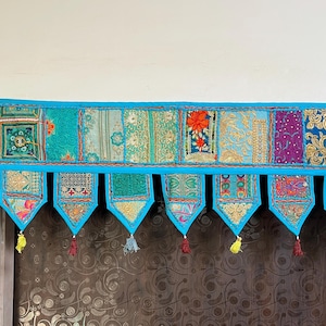 Puede incluir: Una vibrante cenefa textil de patchwork con un borde turquesa. La cenefa presenta una serie de paneles rectangulares en varios patrones y colores, incluyendo turquesa, dorado y morado. Cada panel tiene un colgante triangular decorativo con borlas.