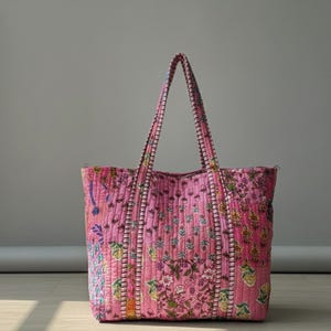 Könnte beinhalten: Eine gesteppte Tragetasche in Rosa mit floralem Muster. Die Tasche hat zwei lange Henkel und besteht aus einem weichen, strukturierten Stoff. Das florale Design umfasst grüne, gelbe und blaue Akzente.