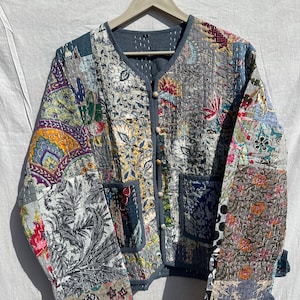 May include: A patchwork jacket with a V-neck and button fastening. The jacket displays a variety of floral and paisley patterns in shades of grey, blue, yellow, and pink. It has two pockets and is hanging on a wooden hanger.