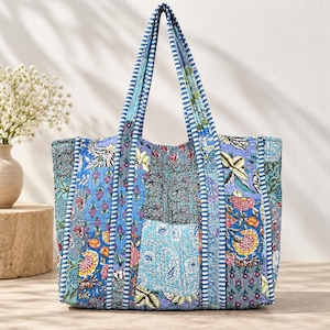 Puede incluir: Un bolso tote acolchado con un diseño de patchwork que presenta varios estampados florales y de cachemira en tonos azul, rosa y amarillo. El bolso tiene dos asas largas con un borde a rayas azul y blanco a juego con el ribete vertical.
