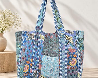 May include: A quilted tote bag featuring a patchwork design with various blue, pink, and yellow floral and paisley patterns. The bag has two long shoulder straps with a blue and white striped trim that matches the vertical piping on the bag.