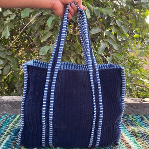 May include: A navy blue tote bag with a corduroy texture. The bag has blue and white striped handles and trim. The bag is held up against a backdrop of green foliage.