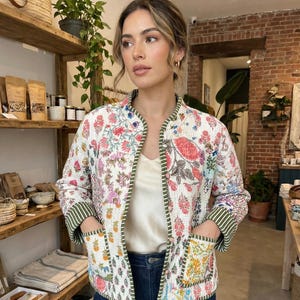 Pode incluir: Casaco acolchoado branco com um vibrante padrão floral em rosa, vermelho e azul. O casaco tem acabamentos verdes e punhos listrados, com bolsos com desenhos botânicos coloridos. Usado sobre um top creme e jeans azuis.