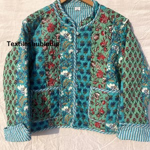 May include: A quilted jacket with a floral print in shades of blue, green, and red. The jacket has a mandarin collar, a button closure, and two pockets. The fabric is a cotton blend.