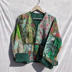 Puede incluir: Chaqueta patchwork con cuello en V y cierre de botones. La chaqueta presenta una variedad de estampados florales y botánicos en tonos verdes, rosas y azules. La chaqueta está colgada en una percha de madera sobre un fondo blanco.