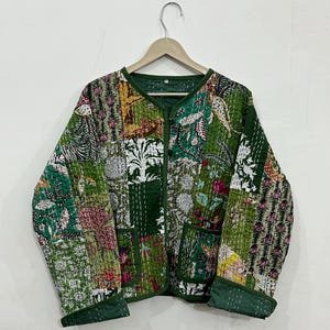 Puede incluir: Chaqueta verde de patchwork con estampados florales y geométricos. La chaqueta tiene cuello en V, dos bolsillos y ribete verde oscuro. La chaqueta está colgada de una percha de madera.