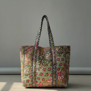 Peut inclure: Un grand sac fourre-tout matelassé avec un motif floral dans les tons de vert, rose et olive. Le sac est doté de poignées et de bordures rayées noires et blanches. Le sac est fait d'un tissu doux et matelassé.