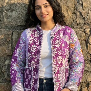 May include: A quilted jacket in shades of purple with a floral pattern. The jacket has a button-up front, striped trim, and is worn over a white shirt and blue jeans. The background is a stone wall.