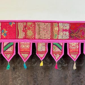 May include: A bright pink textile valance with detailed embroidered panels and decorative tassels. The valance has a patchwork design with gold and colourful thread details. The bottom edge has pendant shapes with tassels in various colours.