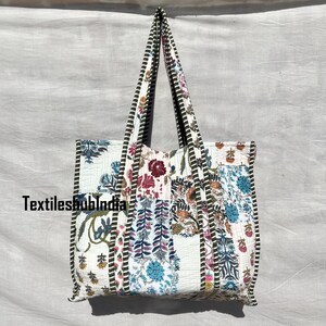 Könnte beinhalten: Eine weiße Baumwoll-Tragetasche mit Patchwork-Design, das florale und geometrische Muster in Blautönen, Grün, Rosa und Lila zeigt. Die Tasche hat einen schwarz-weißen Streifenbesatz und zwei lange Henkel.
