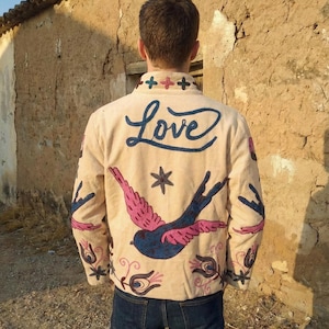 May include: Beige jacket with embroidered design. The back features the word "Love" in blue script, a blue and pink bird, and floral and star motifs. The jacket has a button-up front and long sleeves.