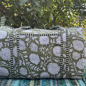 May include: A green and white floral print duffel bag with black and white striped accents. The bag has two handles and a rectangular shape. The floral pattern features large white flowers and leaves on a green background.