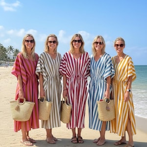 May include: Five women wearing striped caftans in shades of pink, olive, red, blue, and yellow stand on a sandy beach. Each person carries a woven straw tote bag. The caftans have a V-neck and are knee-length.