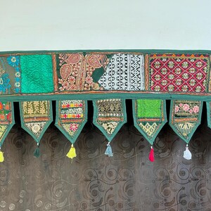 May include: A decorative green textile valance with a patchwork design. The valance features various patterns, including floral and geometric motifs, in shades of green, red, and gold. Each section is bordered with a green fabric and adorned with colorful tassels.