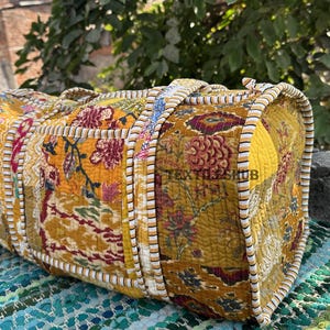 May include: A quilted duffel bag with a floral pattern in shades of yellow, red, and brown. The bag has a rectangular shape with rounded edges and a striped trim. The bag is sitting on a blue and green woven mat.