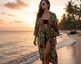 Może przedstawiać: Kobieta w zielonym kimono z kwiatowym nadrukiem i wiązanym pasem spaceruje po piaszczystej plaży o zachodzie słońca. Kimono ma czerwone i różowe wzory kwiatowe. Ma na sobie okulary przeciwsłoneczne i czarny top od bikini.