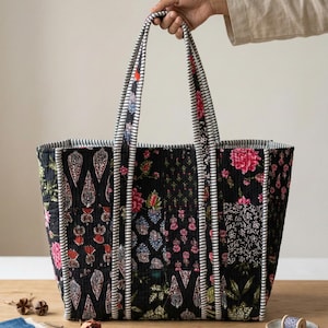 Op de afbeelding: Een grote, gewatteerde draagtas met een zwarte achtergrond en kleurrijke bloempatronen. De tas heeft stevige handvatten met een gestreept ontwerp. De tas wordt door een persoon vastgehouden. De tas staat op een houten oppervlak.
