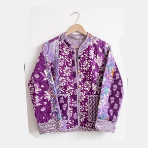 May include: A quilted jacket in shades of purple and white, featuring a floral pattern. The jacket has a round collar, long sleeves, and a button-up front. The jacket is hanging on a wooden hanger.