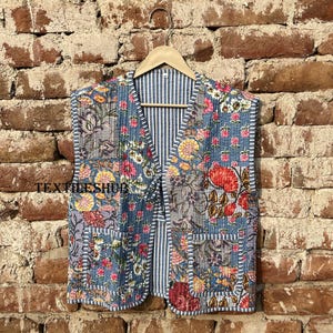 May include: A quilted vest with a floral patchwork design in shades of blue, pink, yellow, and red. The vest has a striped lining and two front pockets. The vest is hanging on a wooden hanger against a brick wall.