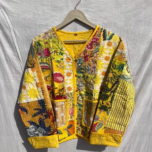 May include: A patchwork jacket with a V-neck and button closure. The jacket features a variety of floral and geometric patterns in yellow, orange, and green. It has two front pockets and is lined with yellow fabric.
