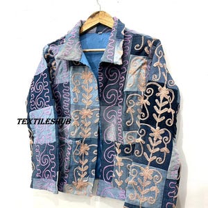 May include: A patchwork denim jacket with pink and light blue floral and swirl embroidery. The jacket has a collared neckline, pockets, and the text "TEXTILESHUB". It is suspended from a wooden hanger.