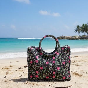May include: A black tote bag featuring a floral design of pink flowers and green foliage. The bag has a circular handle and is placed on a sandy beach with the sea in the background. The edges of the bag are trimmed with a black and white striped pattern.