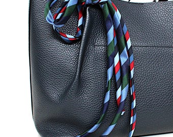 May include: A dark navy blue tote bag with a textured surface and two shoulder straps. A long, thin scarf featuring a diagonal striped pattern in light blue, dark blue, red, and dark green is tied around the top handle of the bag.