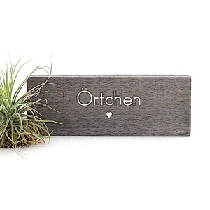 K&ouml;nnte beinhalten: Ein graues Holzschild mit dem Wort "&Ouml;rtchen" in wei&szlig;er, stilisierter Schrift und einem kleinen Herzsymbol. Links vom Schild befindet sich eine gr&uuml;ne Luftpflanze. Das Schild ist ein dekorativer Gegenstand.