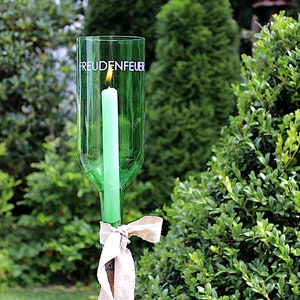May include: A green glass bottle candle holder with the word "FREUDENFEUER" printed on it. A lit green candle is inside the bottle. The holder is tied with a beige ribbon. The background is a green garden.