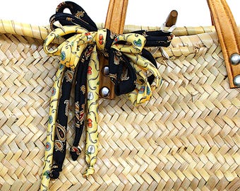 May include: A woven straw basket bag featuring tan leather handles attached with silver-tone metal rivets. A decorative bow made of black and yellow fabric ribbons with a paisley and fruit print is tied around one of the handles on the basket.