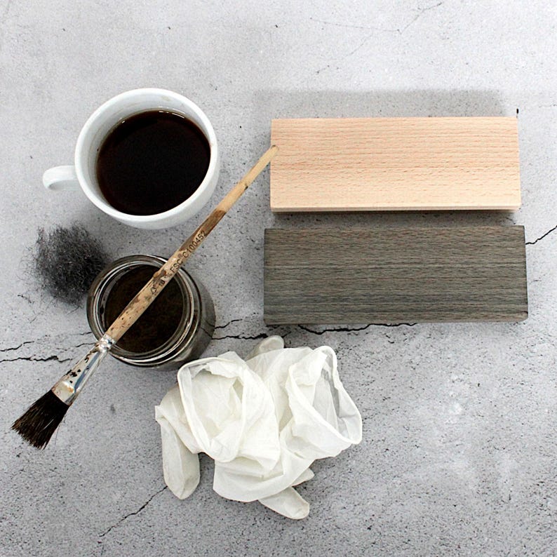 K&ouml;nnte beinhalten: Draufsicht auf Holzbearbeitungszubeh&ouml;r. Eine wei&szlig;e Kaffeetasse, ein Farbtopf, ein Pinsel, zwei Holzbl&ouml;cke, Stahlwolle und ein Paar wei&szlig;er Handschuhe sind auf einer grauen Oberfl&auml;che angeordnet. Ein Holzblock ist naturbelassen, der andere grau gebeizt.