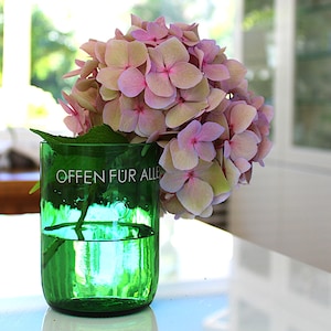 Könnte beinhalten: Eine grüne Glasvase mit dem Text "OFFEN FÜR ALLE" enthält einen Strauß rosa und hellgrüner Hortensien. Die Vase ist mit Wasser gefüllt und steht auf einer weißen Oberfläche, mit einem hellen Fenster im Hintergrund.