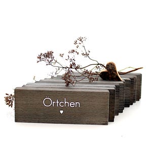 K&ouml;nnte beinhalten: Dekorative Holzpr&auml;sentation mit grauen rechteckigen Bl&ouml;cken. Das Wort "&Ouml;rtchen" wird in Wei&szlig; mit einem kleinen Herzsymbol angezeigt. Ein Trockenblumenarrangement und ein herzf&ouml;rmiges Ornament liegen oben auf den Bl&ouml;cken.