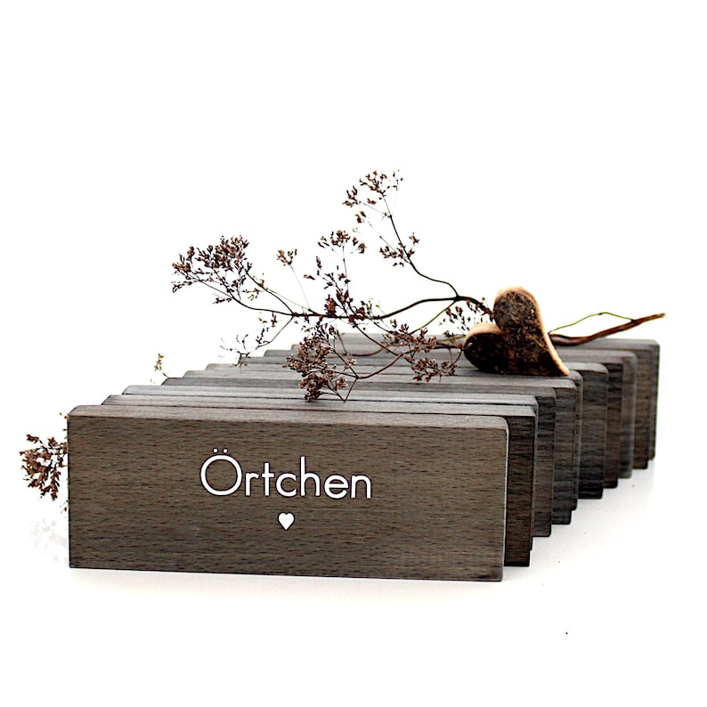 K&ouml;nnte beinhalten: Dekorative Holzpr&auml;sentation mit grauen rechteckigen Bl&ouml;cken. Das Wort "&Ouml;rtchen" wird in Wei&szlig; mit einem kleinen Herzsymbol angezeigt. Ein Trockenblumenarrangement und ein herzf&ouml;rmiges Ornament liegen oben auf den Bl&ouml;cken.