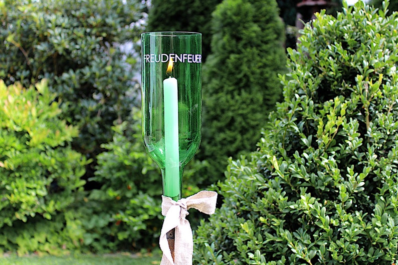 May include: A green glass bottle candle holder with the word "FREUDENFEUER" printed on it. A lit green candle is inside the bottle. The holder is tied with a beige ribbon. The background is a green garden.