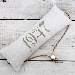 Könnte beinhalten: Ein rechteckiges, helles Leinenkissen mit der Zahl "1947" in verblasstem Olivgrün aufgedruckt. Eine braune Lederschnur mit einem goldfarbenen Knopf ist befestigt. Das Kissen liegt auf einer weißen Holzoberfläche.