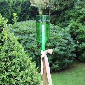 May include: A green glass bottle candle holder with the word "FREUDENFEE" printed on it. A white candle is inside the bottle. The holder is tied with a beige ribbon and mounted on a wooden stake, set in a garden.