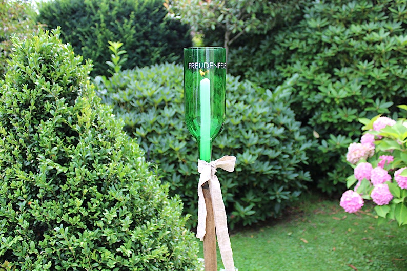 May include: A green glass bottle candle holder with the word "FREUDENFEE" printed on it. A white candle is inside the bottle. The holder is tied with a beige ribbon and mounted on a wooden stake, set in a garden.